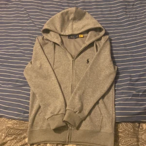 Grå hoodie från Ralph Lauren - Säljer en stilren grå hoodie från Ralph Lauren med dragkedja och huva. Tröjan har långa ärmar och en liten logotyp på bröstet. Perfekt för en avslappnad look.