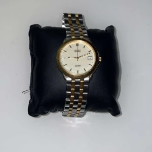 Tvåfärgad klocka från Seiko - Elegant Seiko-klocka med tvåfärgad länkarmband i silver och guld. Urtavlan är vit med datumvisning och guldfärgade markeringar. Perfekt för en stilren look.