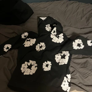 Svart hoodie med blommönster från Denim Tears - Säljer en svart hoodie från Denim Tears med ett unikt vitt blommönster. Hoodien har en oversized passform med huva och långa ärmar. Perfekt för en stilren och avslappnad look.