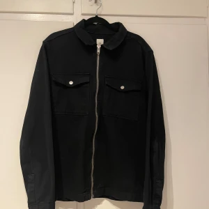 Svart jacka från River Island - Snygg svart jacka från River Island med dragkedja och två bröstfickor med knappar. Perfekt för en stilren look.