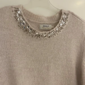 Beige stickad tröja från ONLY - Säljer en elegant beige stickad tröja från ONLY med glittrande pärldetaljer runt halsringningen. Tröjan har långa ärmar och ribbade kanter som ger en stilren look. Perfekt för att addera lite glamour till vardagen.