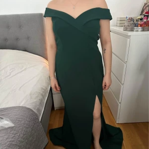 Mörkgrön långklänning från Goddiva - Elegant mörkgrön långklänning från Goddiva med offshoulder-design och en stilfull slits framtill. Klänningen har en dragkedja i ryggen för enkel påklädning. Perfekt för speciella tillfällen.