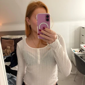 Vit ribbad tröja från Gina Tricot - Säljer en vit ribbad tröja från Gina Tricot med knappar framtill. Tröjan har långa ärmar och en croppad design med vågig nederkant. Perfekt för en stilren look! Supermjuk och endast använd en gång. 