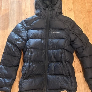 Svart dunjacka från Peak Performance - Säljer en svart dunjacka från Peak Performance. Jackan har en dragkedja framtill och en huva för extra värme. Perfekt för kalla dagar med sin pufferdesign.