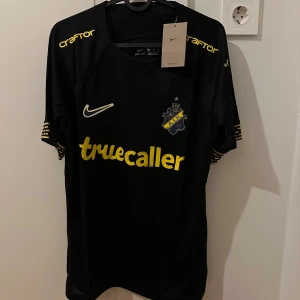 AIK tröja - Säljer en svart fotbollströja från Nike med gula detaljer och tryck från AIK och Truecaller. Tröjan har korta ärmar och är perfekt för fotbollsfans. Märket SVEA syns på baksidan. En stilren design för den hängivne supportern. Produkten är helt ny och oanvänd!