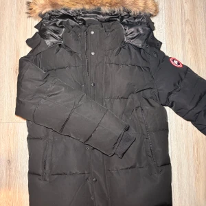 Svart dunjacka från Canada Goose - Säljer en svart dunjacka från Canada Goose med pälsdetaljer på huvan. Jackan har en broderad logotyp på ärmen och är perfekt för kalla vinterdagar. Den är långärmad och har en stilren design med knappar och dragkedja framtill.