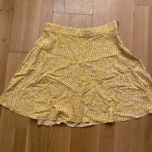 Gul blommig kjol från NA-KD - Säljer en söt gul kjol med vitt blommönster från NA-KD. Kjolen har en avslappnad passform och är perfekt för varma dagar. Den är gjord i ett lätt och bekvämt material.