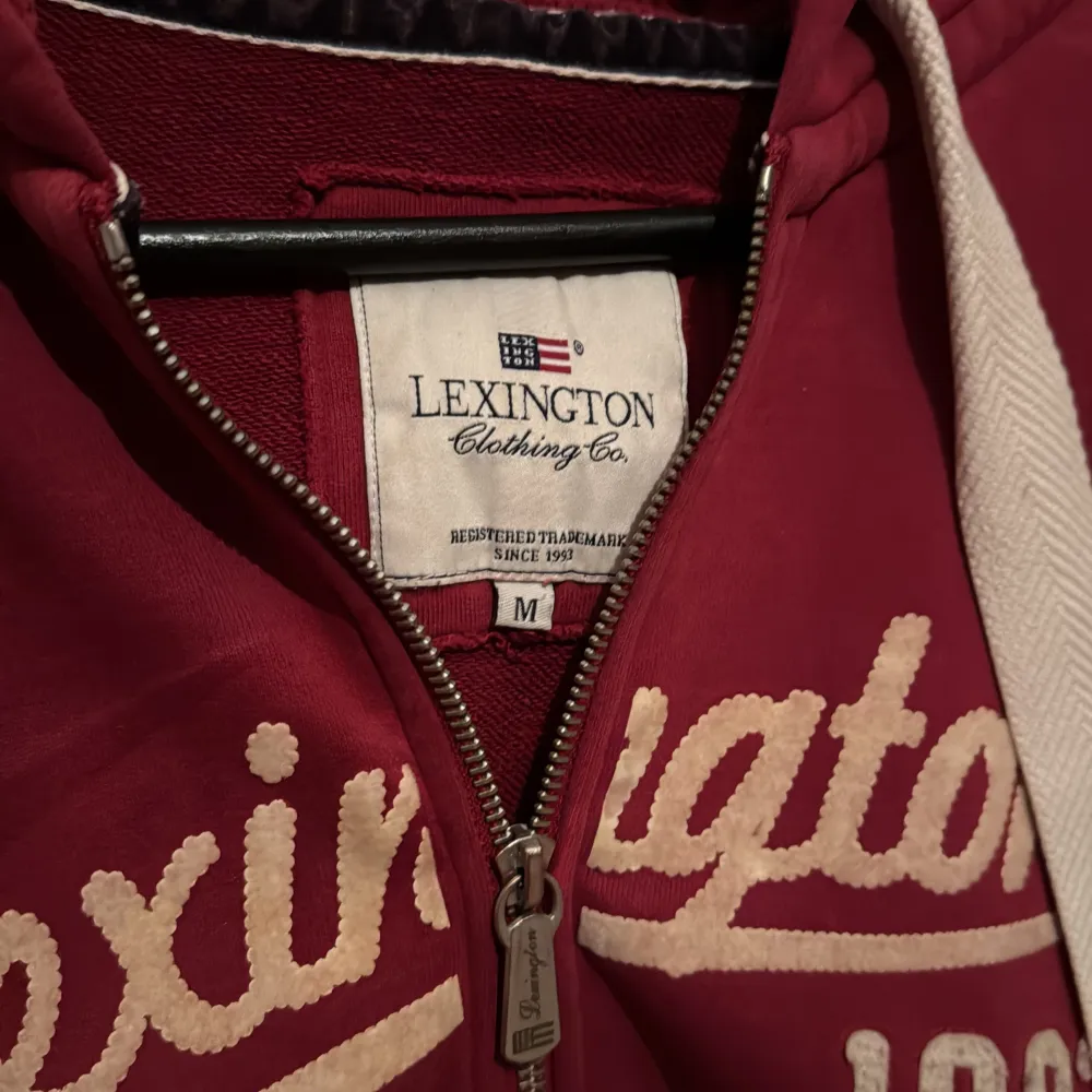 Säljer en snygg röd hoodie från Lexington med dragkedja och vit text 'Lexington 1993' på framsidan. Den har en justerbar huva med vita snören och praktiska fickor framtill. Perfekt för en avslappnad stil.. Neuletakit & Villapaidat.