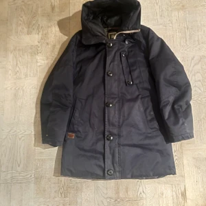 Svart parkajacka från G-Star RAW - Säljer en stilren svart parkajacka från G-Star RAW. Jackan har en huva och knäppning med både dragkedja och knappar. Perfekt för kyligare dagar med sin långa design och praktiska fickor.