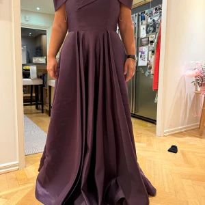 Mörklila långklänning från Bașaǧlu - Elegant mörklila långklänning från Bașaǧlu med offshoulder-design och smala axelband. Klänningen har en vacker omlott-detalj framtill och är tillverkad i ett glansigt material som ger en lyxig känsla. Perfekt för speciella tillfällen ex bal, bröllop, förlovnings fest… Använd en gång på ett bröllop. Den är ca storlek 40-42, dragkedjan har gått sönder men går jättebra att dra upp utan och lätt att byta 