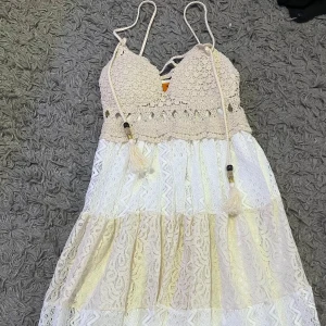 Beige och vit klänning med spetsdetaljer - Charmig klänning i beige och vitt med vackra spetsdetaljer och justerbara axelband. Den har en bohemisk känsla med virkade partier och tofsar som ger en unik touch. Strandklänning som är köpt i Spanien. Ovandel med vaddering. Om du har frågor, vänligen kontakta mig på engelska.🥰🥰Ej använd en enda gång.