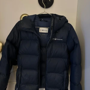 Mörkblå dunjacka från Sail Racing - Säljer en mörkblå dunjacka från Sail Racing. Jackan har en pufferdesign med dragkedja och en praktisk huva. Perfekt för kyliga dagar.