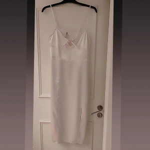 Vit klänning från H&M - Elegant vit klänning från H&M med smala axelband och en söt knytdetalj framtill. Klänningen har en slits som ger en stilren look. Perfekt för sommarens festligheter.