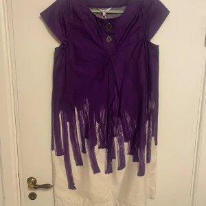 Lila och vit klänning från Signal - Jättefin med ett skärp! Unik lila och vit klänning från Signal med korta ärmar och två dekorativa knappar framtill. Den har en kreativ design med ett penseldragsmönster som ger en konstnärlig touch. Perfekt för dig som vill sticka ut!