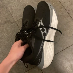 Svarta löparskor från Saucony - Säljer ett par svarta löparskor från Saucony med vit sula och snörning. Skorna har en modern design med svarta detaljer och är perfekta för löpning.