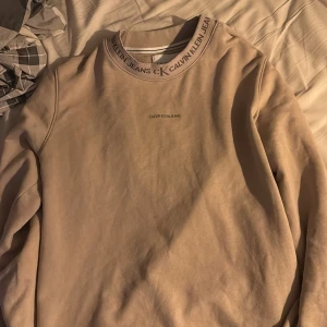 Beige sweatshirt från Calvin Klein Jeans - Säljer en stilren beige sweatshirt från Calvin Klein Jeans. Tröjan har en rund halsringning med logotypdetaljer och är långärmad. Perfekt för en avslappnad look.