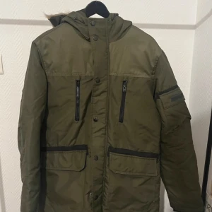 Vinterjacka från Jack & Jones - Snygg grön parkajacka från Jack & Jones med huva och fuskpälsdetalj. Jackan har flera praktiska fickor med dragkedja och knappar. Perfekt för kyliga dagar. Extremt varm och oanvänd, mer eller mindre ny. Kan gå med på nån deal, skicka iväg ett pris/fråga!