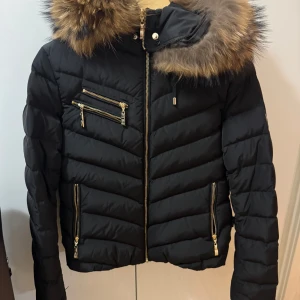 Svart dunjacka från Hollies - Snygg svart dunjacka från Hollies med pälskantad huva och leopardmönstrat foder. Jackan har dragkedja och flera praktiska fickor med gulddetaljer. Perfekt för kyliga dagar. Knappt använd 1 vinter utan fått hänga i garderoben. 