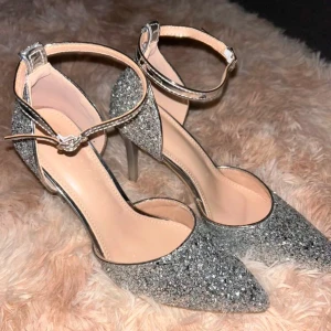 Glittriga silverpumps med ankelrem - Snygga glittriga pumps i silver med spetsig tå och elegant ankelrem. Perfekta för en festlig kväll med hög klack för extra stil.  Oanvända, storlek 38 