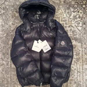 Svart dunjacka från Moncler - Säljer en snygg och varm marinblå dunjacka från Moncler. Jackan har en huva och dragkedja framtill. Den är perfekt för kalla vinterdagar och har en ficka med knapp på ärmen med Moncler-loggan. Jackan är i nyskick och redo att hålla dig varm och stilren i vinter. Kvitto och tags finns. Idag är nypriset nästan det dubbla så du får en bra jacka för ett bra pris.