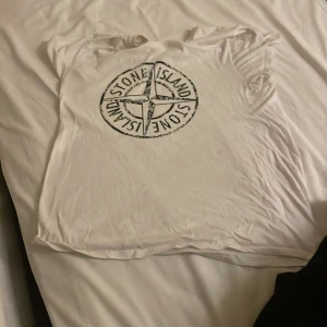 Vit t-shirt från Stone Island - Säljer en vit t-shirt från Stone Island med deras ikoniska logga tryckt på framsidan. T-shirten är i bomull och har korta ärmar. Perfekt för en casual look eller för att visa upp ditt favoritmärke. Den är i välanvänt skick men fortfarande snygg och bekväm.