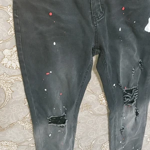 Svarta jeans med slitningar och färgstänk - Säljer ett par svarta jeans med coola slitningar och färgstänk i rött och vitt. Jeansen har en tapered passform och är perfekta för en avslappnad och trendig look. De är i bra skick och redo för en ny ägare!