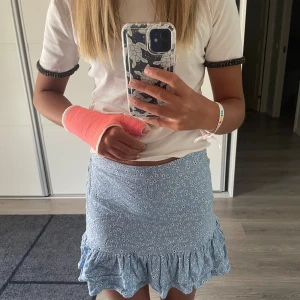 Blå blommig kjol med volang - Säljer en superfin blå kjol med blommigt mönster. Kjolen är kort och har en söt volangdetalj längst ner. Perfekt för sommaren eller en fest! Den är i nyskick och passar perfekt till en vit topp eller t-shirt. Materialet känns lätt och luftigt, vilket gör den bekväm att bära hela dagen.