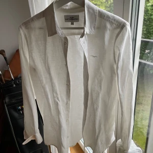 Vit linneskjorta från Hampton Republic - Säljer en snygg och stilren vit linneskjorta från Hampton Republic. Skjortan har långa ärmar och är tillverkad i ett lätt och luftigt linnematerial, perfekt för varma dagar. Den har knappar framtill och en liten broderad logga på bröstet. Passar både till vardags och fest! Varsamt använd och har inga tecken på slitage.