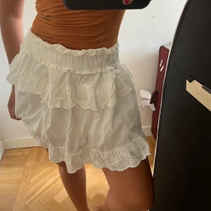 Vit volangkjol - Säljer en superfin vit kjol med volanger. Den är kort och har en söt, romantisk stil med spetsdetaljer. Perfekt för sommaren eller en festlig tillställning. Kjolen är i nyskick och passar perfekt till en avslappnad eller uppklädd outfit.