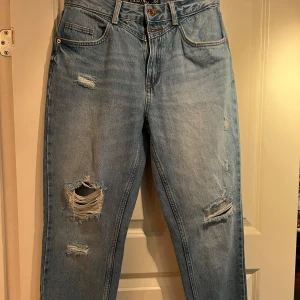 Blå mom jeans från Amisu - Säljer ett par snygga blå mom jeans från Amisu i storlek 38. De har en cool sliten look med flera revor på både fram- och baksidan. Perfekta för en avslappnad och trendig stil. De är högmidjade och har en knapp och dragkedja framtill. Passar perfekt till vardags eller en chill utekväll. 🥰