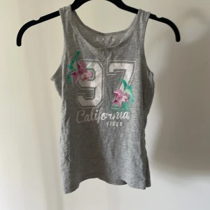 Grå ärmlös topp med tryck från Reaction - Säljer en grå ärmlös topp från Reaction med trycket '97 California Vibes' och blommor i rosa och grönt. Perfekt för sommardagar eller träning. Toppen är i bomull och väldigt bekväm att ha på sig. Passar bra till jeans eller shorts! storlek 134-140.