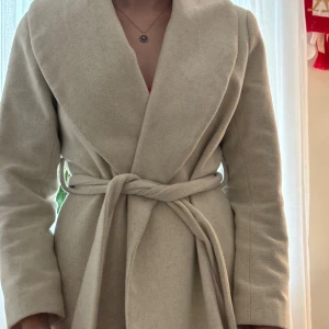 Beige kappa med bälte - Säljer en snygg beige kappa i storlek XS. Den är långärmad och har ett bälte i midjan för en fin passform. Perfekt för höst och vår! Jackan är i bra skick och väldigt bekväm att ha på sig. Passar både till vardags och fest.