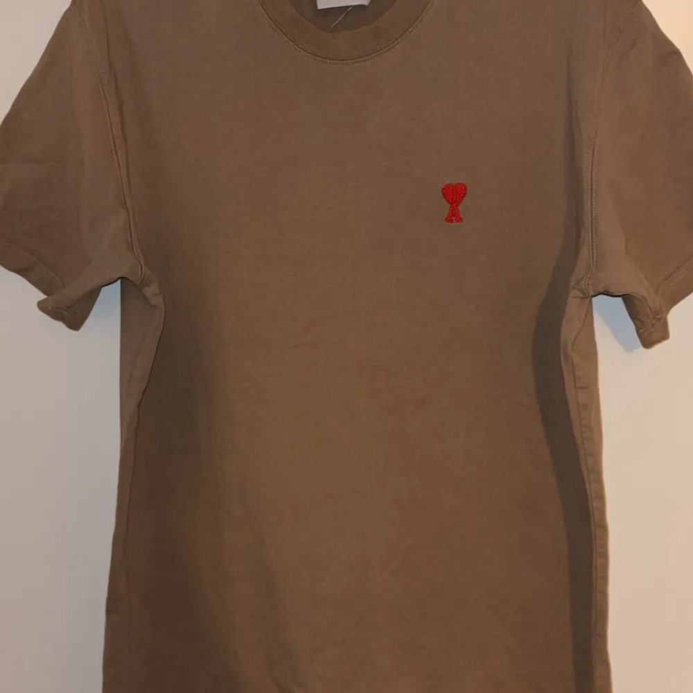 Säljer en snygg beige t-shirt från AMI i storlek L kan passa M den är i väldigt bra skick  bra skick. Den har en liten röd broderad detalj på bröstet som ger en cool touch. Perfekt för vardagsbruk eller en avslappnad dag ute. Passar bra till jeans eller shorts!. T-shirts.