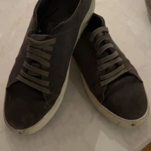 Bruna sneakers i mocka - Arigato Säljer ett par bruna sneakers i mocka med snörning. De har en klassisk design med vit sula och är perfekta för vardagsbruk. Skorna är i bra skick och passar till både jeans och chinos.