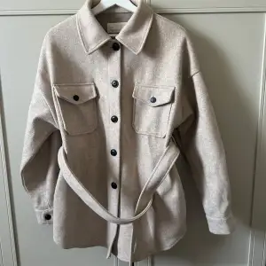 Säljer en snygg beige overshirt från H&M i mycket bra skick. Den har långa ärmar och knappar framtill samt två bröstfickor med knappar. Jackan är i ullmaterial och har ett bälte i midjan för en stilren look. Perfekt för höst och vår!