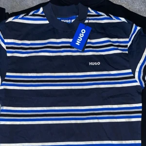 Randig t-shirt från Hugo - Säljer en snygg randig t-shirt från Hugo i blått och vitt. Den har korta ärmar och är perfekt för sommaren. T-shirten är i nytt skick och har en normal passform. Märket 'Hugo' är broderat på bröstet. Perfekt för en avslappnad stil!
