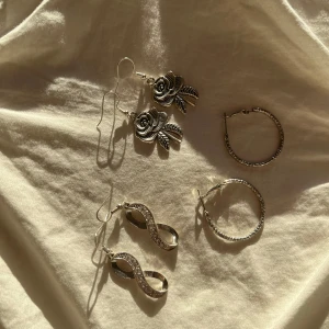 Tre par silverfärgade örhängen med detaljer - Säljer tre par snygga örhängen i silverfärg. 35kr/styck Ett par med rosdesign och detaljerade blad, ett par med en elegant twistad form (som evighetstecknet) med glittriga stenar, och ett par klassiska creoler med en glittrig yta. Perfekta för att piffa upp vilken outfit som helst! 🌟