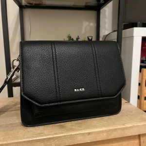 Svart axelväska från NA-KD - Snygg svart axelväska från NA-KD. Väskan har en stilren design med en praktisk dragkedja och en justerbar axelrem. Perfekt för både vardag och fest!