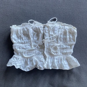 Vit volangprydd tubtopp från Zara - Säljer en superfin vit tubtopp från Zara i storlek S. Toppen har volanger och snörning framtill för en söt och romantisk look. Materialet är linne 