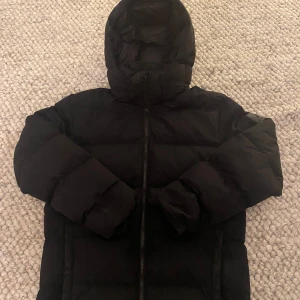 Svart dunjacka från Mckinley - Säljer en svart dunjacka från Mckinley i storlek medium. Jackan är perfekt för kalla vinterdagar med sin tjocka vaddering och huva. Den har en dragkedja framtill och är i mycket bra skick. Värt att notera liten bristning över ena fickan och armhålan men annars perfekt för att hålla sig varm och stilren under vintern! 