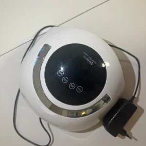 SUNUV nagellampa - Säljer en SUNUV nagellampa i vitt med svart display och silverdetaljer. Perfekt för att härda gelnagellack snabbt och effektivt. Den har flera tidsinställningar och kommer med en strömadapter. Använd för att få salongskvalitet hemma!