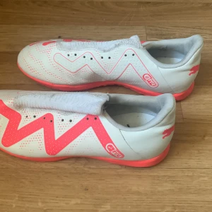 Vita och rosa fotbollsskor från Puma - Säljer ett par vita fotbollsskor från Puma med coola rosa detaljer och mönster. Skorna har en slimmad passform och är perfekta för inomhusfotboll. De är i bra skick och redo för nya äventyr på planen. Storlek passar de flesta.