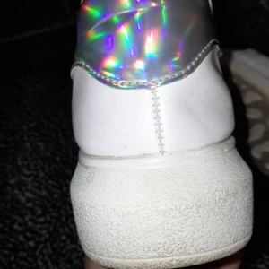 Vita sneakers med holografisk detalj - Säljer ett par vita sneakers i bra skick med en cool holografisk detalj på hälen. De har snörning och en mönstrad sula som ger bra grepp. Perfekta för både vardag och festliga tillfällen!