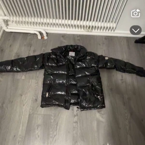 Svart pufferjacka från Moncler - Säljer en snygg svart pufferjacka från Moncler i mycket bra skick. Jackan har en glansig finish och är perfekt för kalla vinterdagar. Den har en dragkedja framtill och en cool Moncler-logga på ärmen. Perfekt för dig som vill hålla stilen i vinter! 🖤