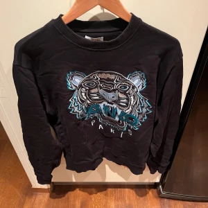 Svart tröja med tigerbroderi från Kenzo - Säljer en svart tröja från Kenzo med ett coolt tigerbroderi på framsidan. Tröjan har långa ärmar och är i bra skick. Perfekt för höst och vinter, och den är både stilren och bekväm. Hör av er vid intresse!