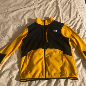 Gul och svart fleecejacka från The North Face - Säljer en snygg gul och svart fleecejacka från The North Face. Den har en dragkedja framtill och en liten bröstficka med dragkedja. Perfekt för höst och vår, och den är i mycket bra skick. Jackan har långa ärmar och är supermjuk och bekväm. Perfekt för utomhusaktiviteter eller bara för att hålla sig varm! Storlek XL men sitter som L