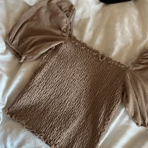 Beige smockad topp - Säljer en superfin beige topp med smockad design och puffiga kortärmar. Perfekt för sommardagar eller en avslappnad vårlook. Toppen har en elastisk passform som gör den bekväm och stilren. Passar både till jeans och kjol!💞