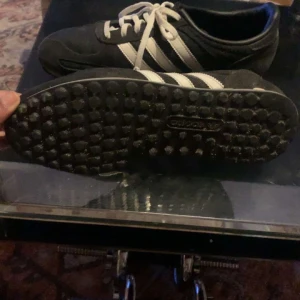 Svarta Adidas LA Trainer sneakers - Säljer ett par klassiska svarta Adidas LA Trainer sneakers i bra skick. De har vita ränder och snörning, med en robust sula perfekt för vardagsbruk. Skorna är gjorda av mocka och syntetmaterial, vilket ger en snygg och hållbar design. Perfekta för både vår och höst!