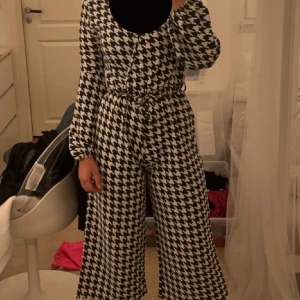 Svartvit mönstrad jumpsuit - Säljer en supercool svartvit jumpsuit med ett iögonfallande rutigt mönster. Den har långa ärmar och vida ben, perfekt för en stilren look. Passar både till vardags och festliga tillfällen. Bekväm och trendig!