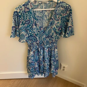 Mönstrad klänning från Zara - Säljer en superfin mönstrad klänning från Zara i blå och lila toner. Den har en v-ringad design med korta ärmar och ett elastiskt band i midjan för en smickrande passform. Perfekt för sommarens alla tillfällen! 🌸